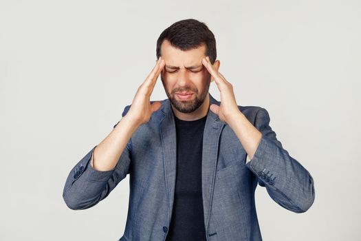 Young businessman man with a beard in a jacket. Hands on head, suffering from headache in despair and under stress due to pain and migraine. Portrait of a man on a gray background.