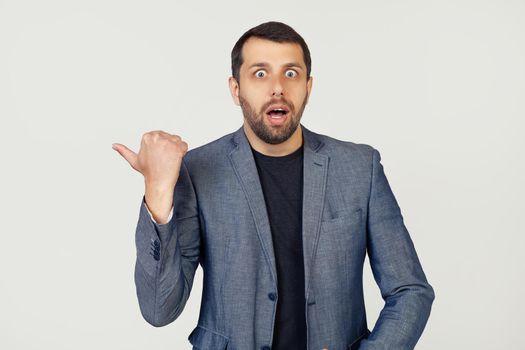 Young businessman man with a beard in a jacket, surprised, pointing finger to the side, with open mouth amazed expression. Portrait of a man on a gray background.