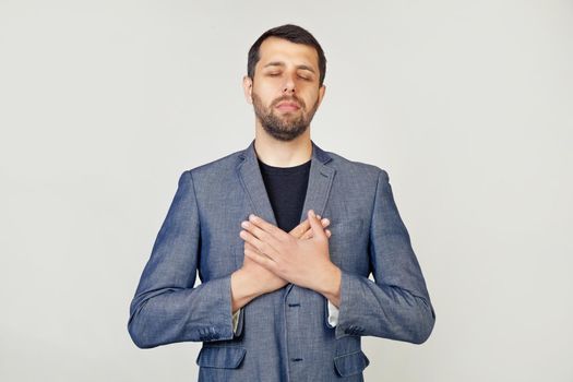 Young businessman with a smiling man with a beard in a jacket, with hands on his chest with closed eyes and a grateful gesture on his face. Health concept. Portrait of a man on a gray background.