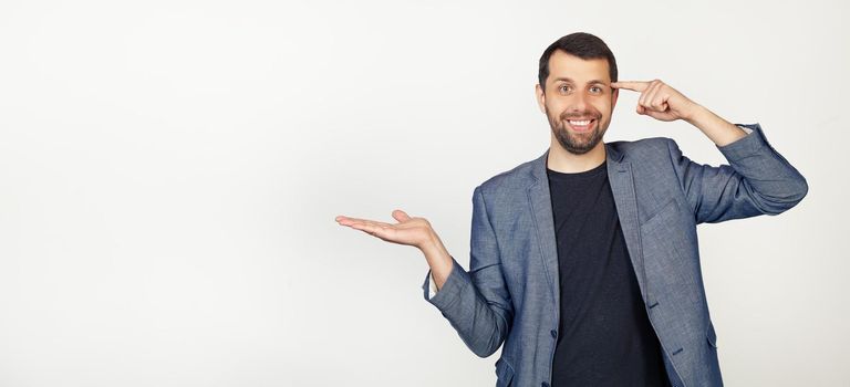 Young businessman man with beard smiling and happy with open palm showing copy space and pointing finger to forehead. I'm thinking about it. Portrait of a man on a gray background.