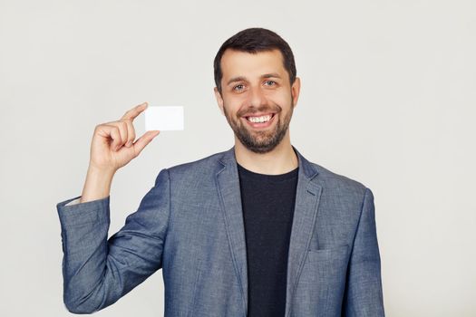 Young businessman man with a beard in a jacket showing a blank sheet of paper. The man smiles and holds in his hand shows a business card. Portrait of a man on a gray background.