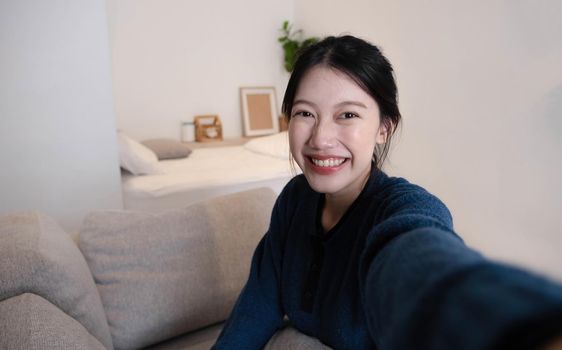 Portrait of Young Asian woman video call with her friend and checking social media by smartphone sitting on couch. Lifestlye concept..