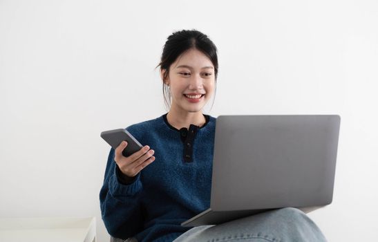 portrait of a happy casual asian woman holding laptop computer and hold smartphone while sitting on a chair over white background.