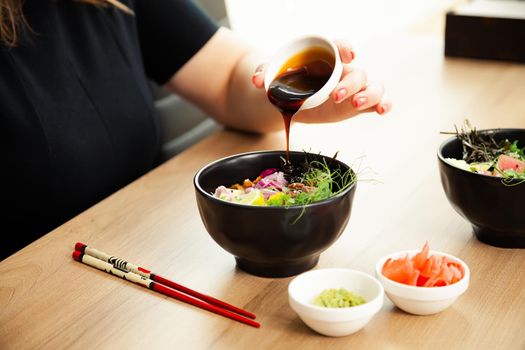 Girl adding teriyaki sauce to Poke salad. Dressing salad with sauce in a restaurant or cafe
