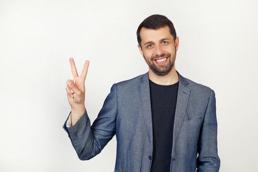 Young businessman man with a beard in a jacket, smiling, looking at the camera, showing thumbs up, making a victory sign. Number two. Portrait of a man on a gray background.