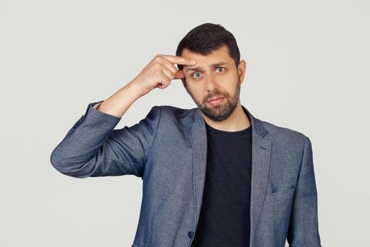 Young businessman man with a beard in a jacket, pointing to an unhappy pimple on his forehead, an ugly acne infection. Acne and skin problems. Portrait of a man on a gray background.