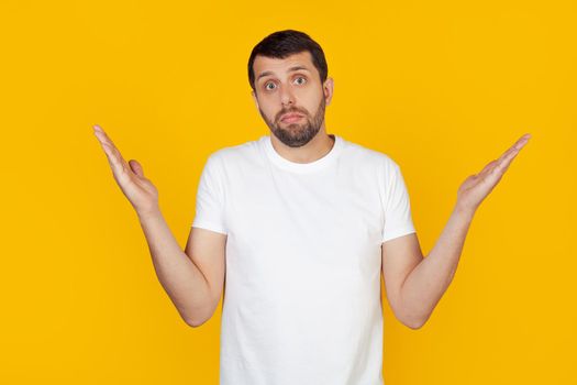 A young man with a beard in a white t-shirt is an ignorant and confused expression with raised arms and hands. Doubt concept. Stands on isolated yellow background.