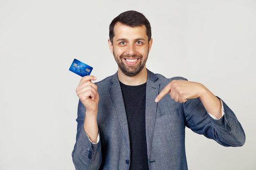 Young businessman man with beard in jacket holding credit card standing on isolated white background with surprised face pointing with finger at himself. Portrait of a man on a gray background.