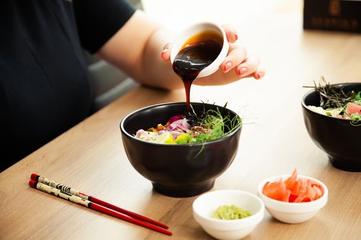 Girl adding teriyaki sauce to Poke salad. Dressing salad with sauce in a restaurant or cafe