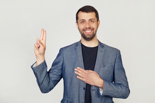 Young businessman man with a beard in a jacket, smiling, swears, putting his hand on his chest and thumbs up, swearing an oath of allegiance. Portrait of a man on a gray background.