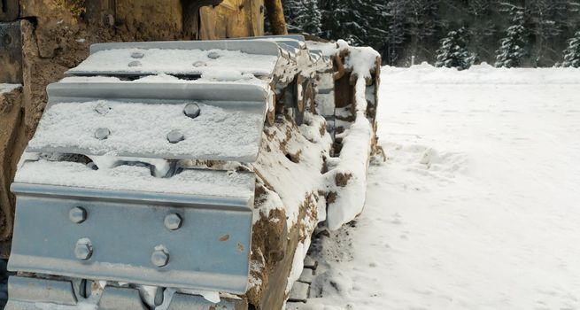 The metal tracks of a bulldozer tractor in winter clears the road from snow. On a frosty day, a yellow bulldozer in winter