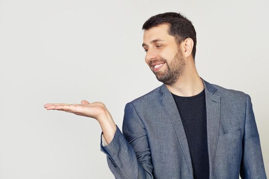 Young man with a beard in a white t-shirt smiling, cheerful presenting and pointing with his palm, looking at his hand. Stands on isolated yellow background.