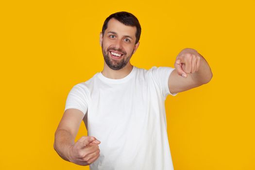 Young man with a beard in a white t-shirt pointing with his fingers at the camera with a happy and funny face. Good energy and vibes. Stands on isolated yellow background.