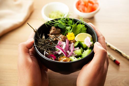 A man holds a Poke salad with beef in a bowl in his hands. Ingredients Beef, Nameko mushrooms, cherry tomatoes, rice, cucumber, red onion, sesame seeds, cilantro, lime. Asian salad concept.