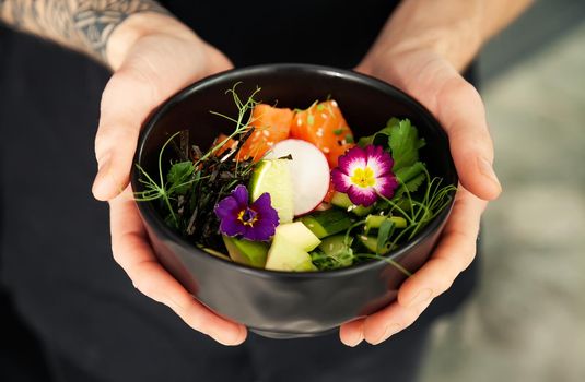 A man holds a bowl of Poke salad with salmon, avocado, cucumber, rice, Tamarind sauce, mango sauce, nori, lime, sesame, cilantro. Asian seafood salad concept