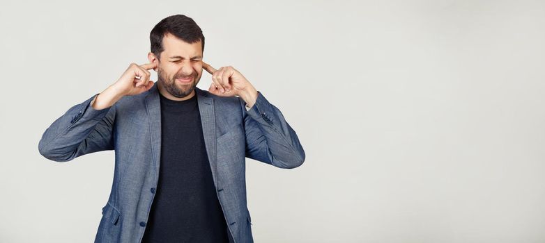 Young businessman man with a beard in a jacket covering his ears with his fingers with an annoyed expression on his face due to the noise of loud music. Deaf concept. man on a gray background.