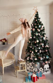 Happy young woman decorates the Christmas tree. A girl in a sweater sets a star on top of a Christmas tree. New year and christmas holiday concept