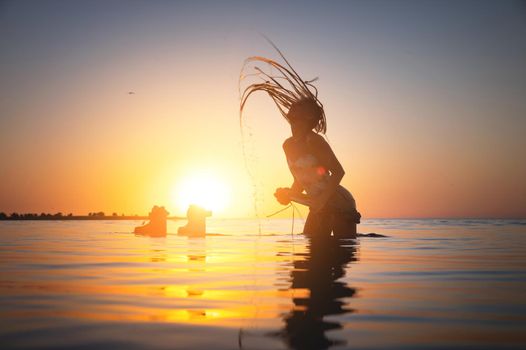 Silhouette frame at sunset. A young female kitesurfer stands up to her feet in sea water next to her floating kiteboard. The girl shakes her wet long hair. Sexy water sports.