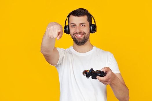 Young man with a beard in a white t-shirt smiling, gamer playing video games with headphones, pointing his finger at the camera and at you, hand sign, positive and confident gesture in front