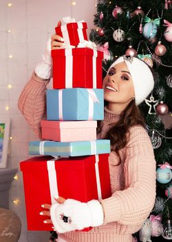 Smiling and happy, young woman in winter clothes holds many wrapped gift boxes in front of her near the Christmas tree. Girl enjoy Christmas gifts, greet New Year and Christmas, holidays concept.