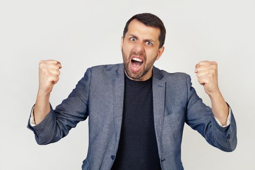 Young businessman man with a beard angry and insane, raising his fists, frustrated and furious, screaming in anger. Rage and aggressive concept. Portrait of a man on a gray background.