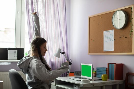 Green screen on laptop. A young girl studies at her desk at home and exercises her hands and arms every now and then. Talented student. Young girl with braids.