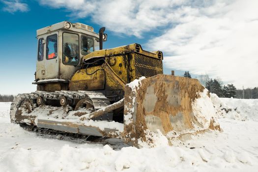 Yellow crawler tractor bulldozer with a bucket works in winter clearing the road from snow. Frosty day with bright sun and beautiful blue sky in the background. Yellow bulldozer in winter
