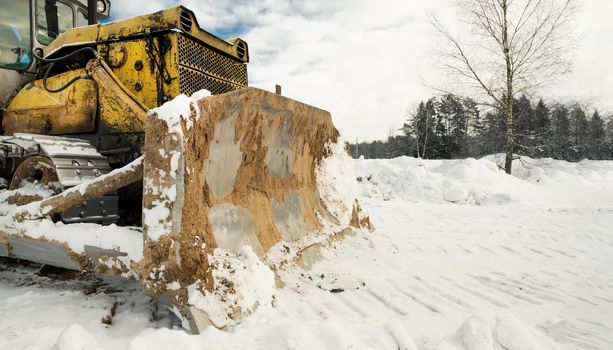 Yellow crawler tractor bulldozer with a bucket works in winter clearing the road from snow. Frosty day with bright sun and beautiful blue sky in the background. Yellow bulldozer in winter