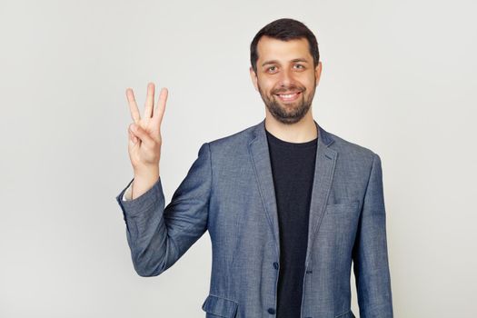 Young businessman with a beard smiles, male businessman in a stylish jacket showing the number three with his fingers, smiling confidently and happy looking at the camera