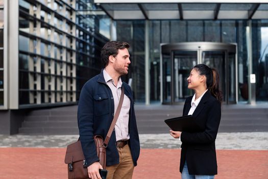 caucasian man and asiatic woman talking about work in front of the entrance to an office building, concept of business and lifestyle, copy space for text