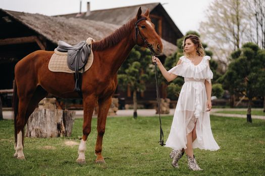 Beautiful girl in a white sundress next to a horse on an old ranch.