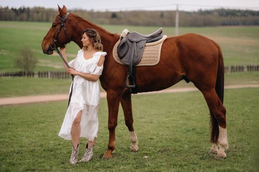 A girl in a white sundress stands next to a brown horse in a field in summer.