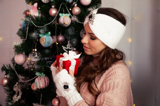 Happy woman holding a gift box on the background of a Christmas tree. Portrait of a happy smiling girl looking at a delivered wrapped box near a decorated fir tree.