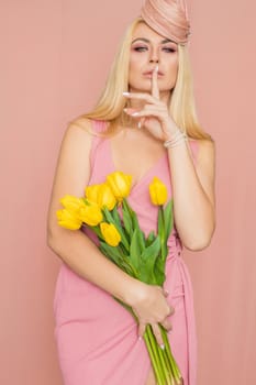 Adult blonde woman in pink dress posing over rosy background. Spring-summer clothing. She is holding bouquet of yellow tulips in her hands. Presents, surprise