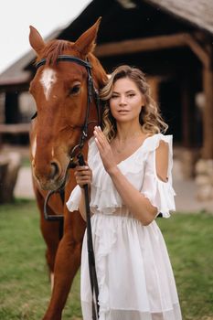 Beautiful girl in a white sundress next to a horse on an old ranch.