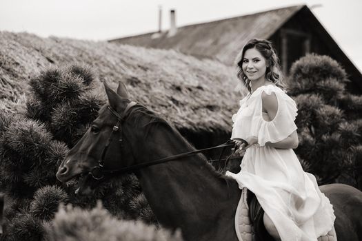A woman in a white sundress riding a horse near a farm. black and white photo.