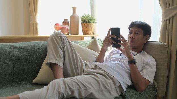 Relaxed man lying on couch in bright living room and using mobile phone for for communicating in social network.