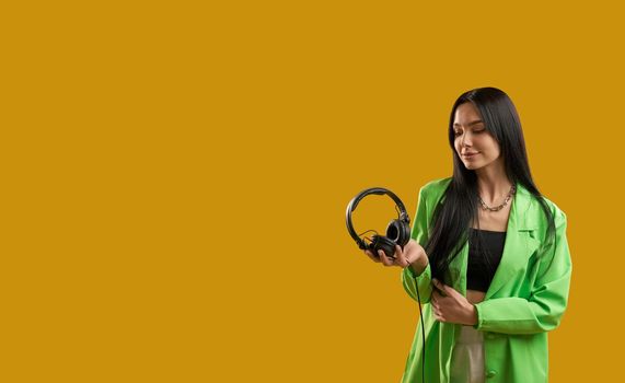 Front view of fashionable girl standing, holding earphones, smiling, Pretty brunette young woman with long hair looking down, enjoying. Isolated on yellow studio background.