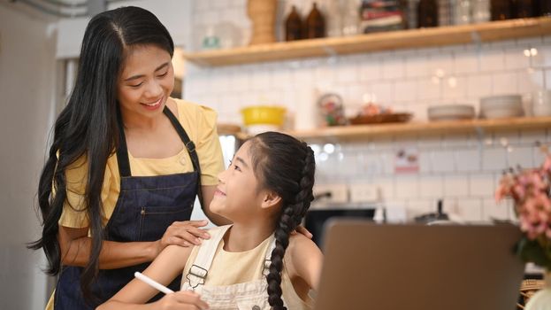 Smiling asian mother helping her little daughter with distance learning virtual distance online class on laptop computer.