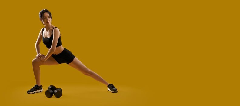 Side view of brunette girl with ponytail stretchig indoors. Slim young pretty female standing, working out, making step, looking forward. Isolated on yellow studio background.