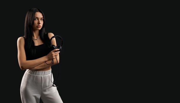 Front view of pretty brunette girl standing, holding earphones. Young beautiful fashionable female with long hair looking forward, thinking. Isolated on black studio background.