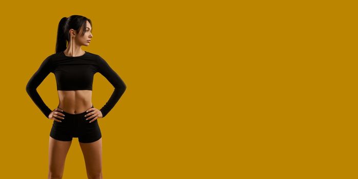 Front view of brunette girl with ponytail working out indoors. Sporty slim young female standing, holding hands on waist, looking aside. Isolated on yellow studio background.