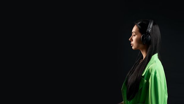 Side view of brunette girl with long hair standing, listening to music. Pretty slim female looking forward with closed eyes, thinking. Isolated on black studio background.