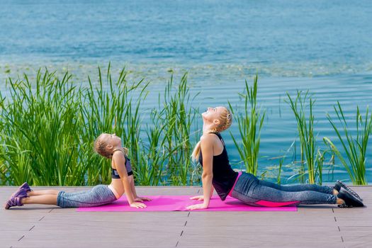 Little girt and woman are doing exercises on the grass at the shore of the lake.