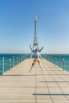 Large model of the Eiffel Tower on the beach. A woman walks along the pier towards the tower, wearing a blue jacket and white jeans