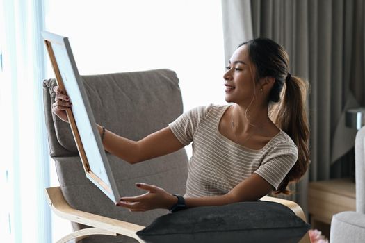 Happy young woman sitting on armchair in living room and looking at her artwork with admiration.