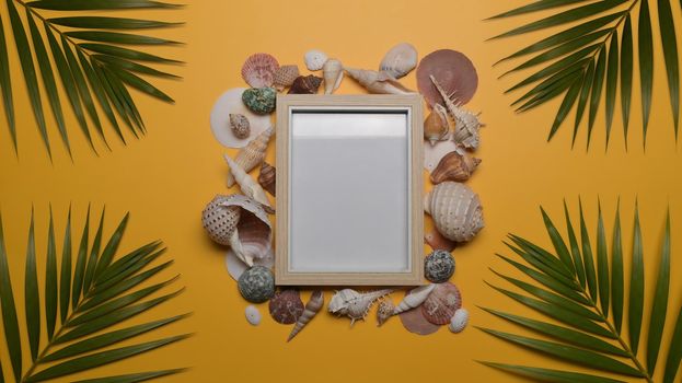 Empty picture frame, seashells and tropical palm leaves on yellow background. Travel, summer and holiday concept.