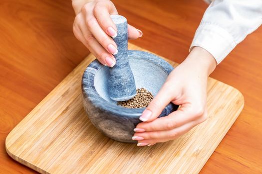 Hands of woman grinding black pepper by pestle in a gray granite mortar on the table.