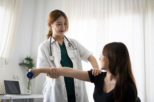 Young woman patient using dumbbells with outstretched arms while getting treatment from physiotherapist.