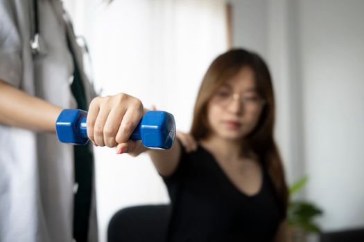 Young woman patient using dumbbells with outstretched arms while training with physiotherapist at clinic.